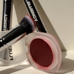 A makeup product shot featuring a round compact of deep berry-red cream blush or eyeshadow, alongside three black makeup brushes with silver ferrules and white 'BLUENCY' branding, arranged on a warm off-white surface with strong sunlight and shadows.
