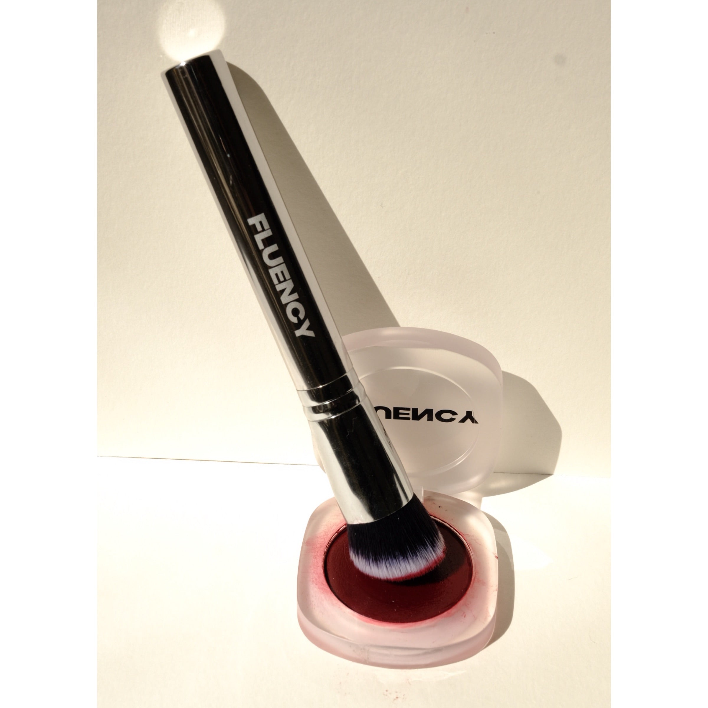 A makeup brush with a black handle and white bristles, branded 'FLUENCY', resting in a clear round case filled with deep red makeup. The case lid is open behind the brush, and the product is photographed on a plain light background with strong direct lighting and shadows.
