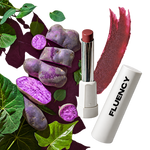 A lipstick tube with its cap removed stands beside a white cylindrical tube printed with the brand name FLUENCY. Purple sweet potato slices and green leaves are arranged around the product, with a pinkish-purple cosmetic smear behind it on a white background.